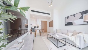 A modern living room with a sofa, coffee table, and dining area. There is a kitchen in the background with a microwave and oven. - StayShort - Luxury 1-Bedroom JVC Apartment with Balcony & Pool (JVC)