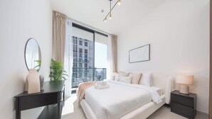 A modern bedroom with a large bed, a bedside table, a nightstand, a mirror, and a view of the city through a large window. - StayShort - Luxury 1-Bedroom JVC Apartment with Balcony & Pool (JVC)
