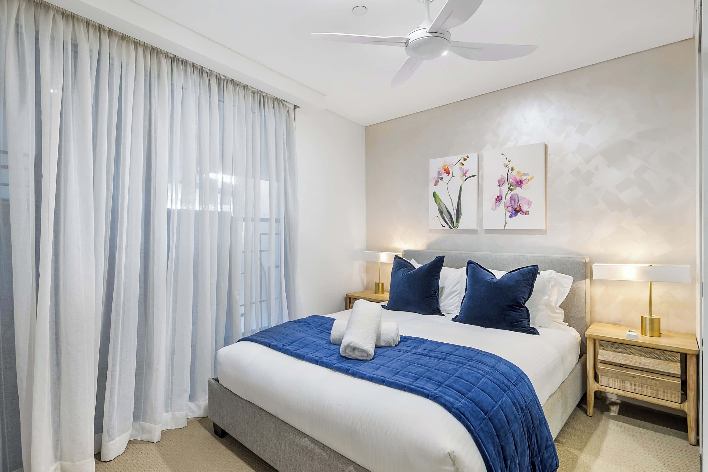A modern hotel room with a large bed, two bedside tables with lamps, a ceiling fan, and sheer curtains.