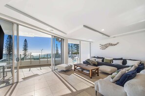 A spacious living area with a large sliding glass door leading to a balcony, a sofa set, a coffee table, and a decorative wall piece. - Oceans 203 Luxury Beachfront Apt (Mooloolaba)