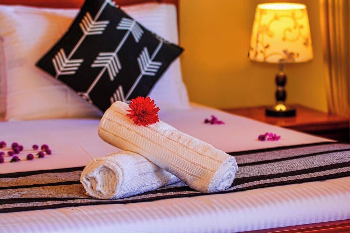 Una cama bien tendida con una toalla enrollada y una flor roja, un cojín blanco y negro y una lámpara de noche.