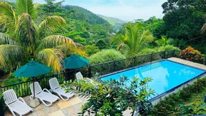 Una piscina con sillas de descanso y sombrillas, rodeada de abundante vegetación y colinas.