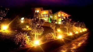 Una casa de dos pisos con balcón, rodeada de caminos y plantas iluminadas.