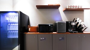 Une petite cuisine équipée d’un réfrigérateur, d’un four à micro-ondes, d’une machine à café et d’une étagère où sont rangées des conserves.