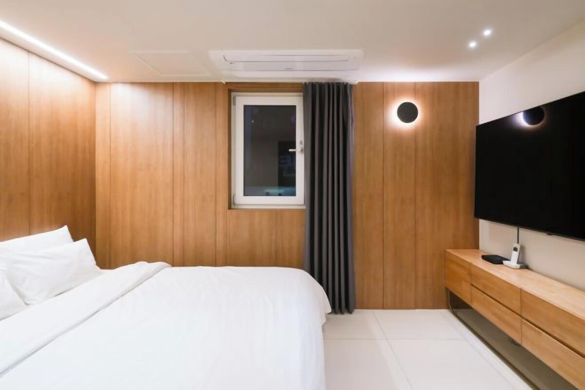 Un dormitorio moderno con pared de paneles de madera, una cama grande con ropa de cama blanca, una ventana con cortinas, un televisor empotrado y una mesita de noche de madera.