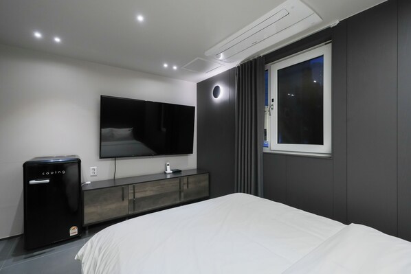 Una habitación de hotel moderna con una cama grande, un televisor de pantalla plana montado en la pared, un mini refrigerador y una ventana con cortinas.