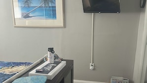 Ein Zimmer mit einem Bett, einem an der Wand montierten Fernseher, einem gerahmten Bild und einer kleinen Holzkiste.