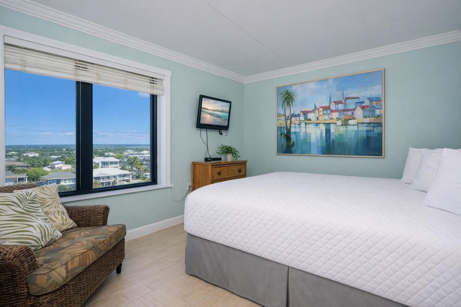A bedroom with a large bed, a view of the city, a painting, and a wicker chair.