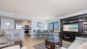 A modern living room with a large TV, comfortable seating, a dining area, and a kitchen in the background. - Island Winds East 501 3 Bedroom Condo (Gulf Shores)