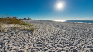 A wide sandy beach with footprints leading to the water, a clear sky, and buildings in the background. - Island Winds East 301 3 Bedroom Condo (Gulf Shores)