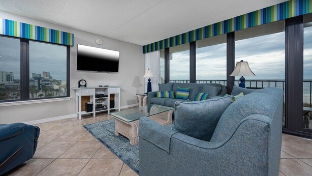 A modern living room with a grey sofa, a coffee table, and a TV on the wall. There is a balcony with a view of the city. - Island Winds East 301 3 Bedroom Condo (Gulf Shores)