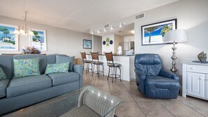 A living room with a sofa, a glass coffee table, a blue leather chair, and a kitchen area with a refrigerator and bar stools. - Island Winds East 301 3 Bedroom Condo (Gulf Shores)