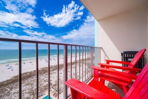 A balcony with a view of a beach, red chairs, and a pool. - Island Winds East 707 2 Bedroom Condo (Gulf Shores)