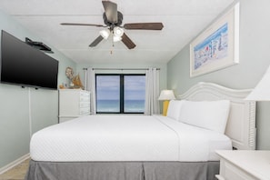 A bedroom with a bed, a ceiling fan, a TV, a dresser, a lamp, and a painting.
