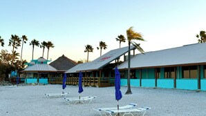 Een gebouw aan het strand met een schuin dak, palmbomen en strandstoelen.