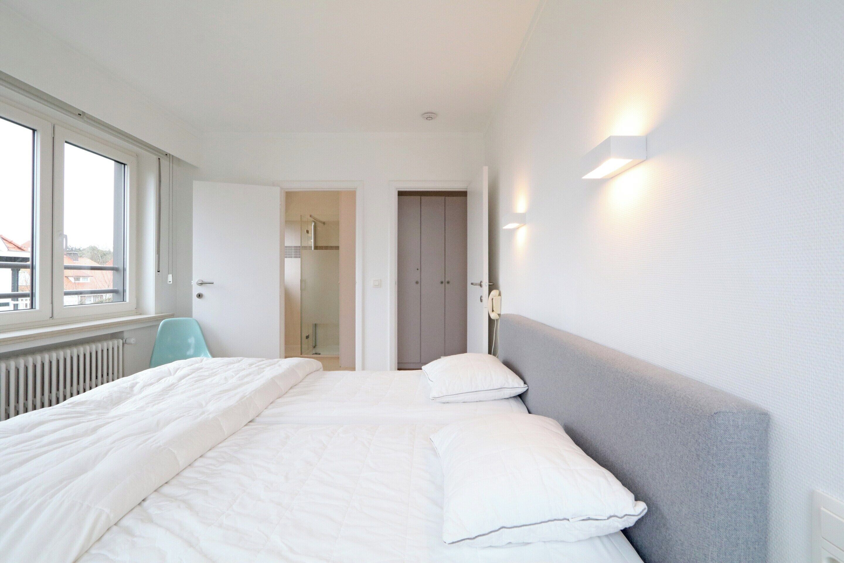 A hotel room with a large bed, a bedside table, a chair, and a view of the city through the window.