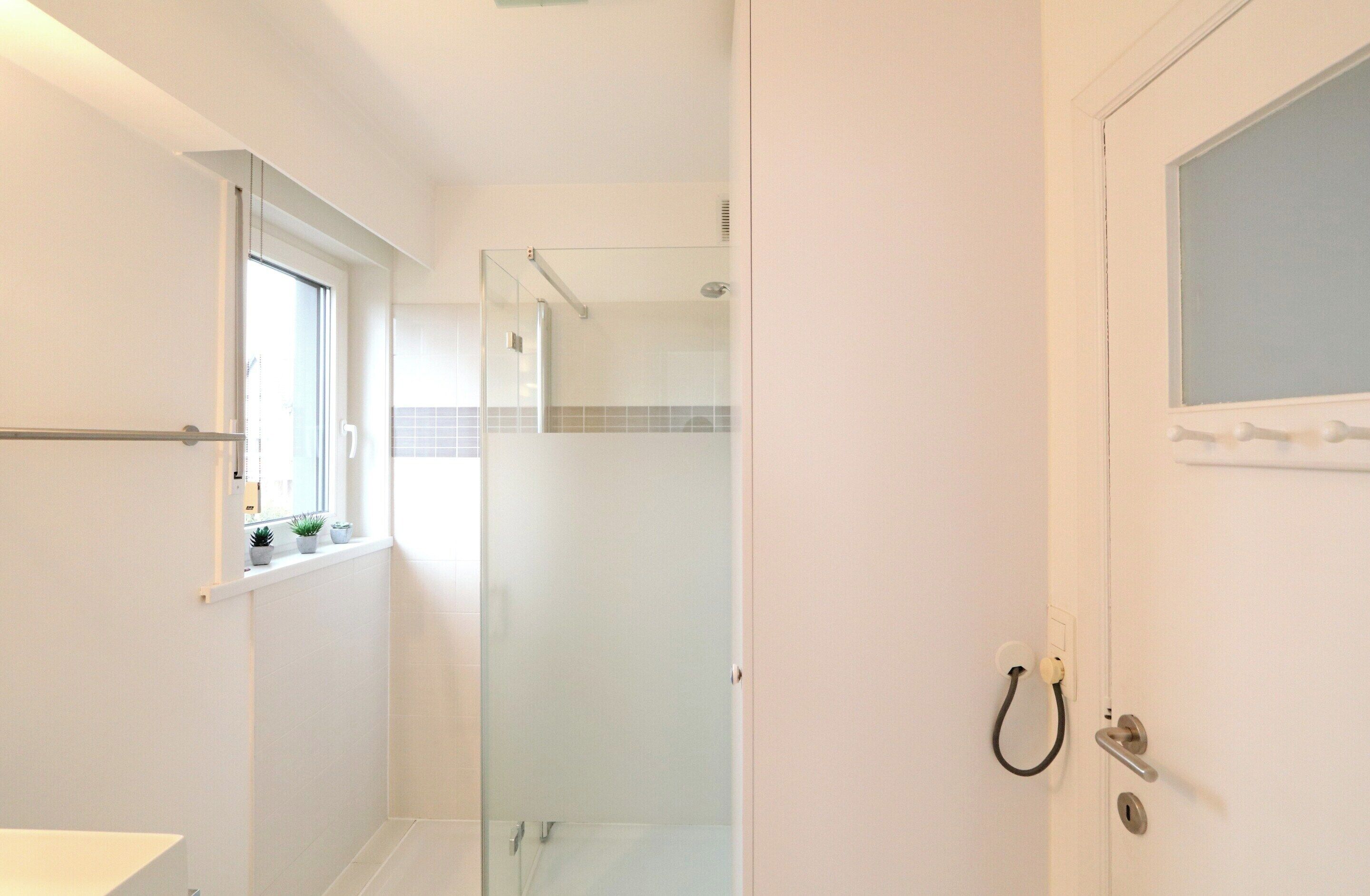 A bathroom with a glass shower enclosure, a window with plants, and a white door with a handle.
