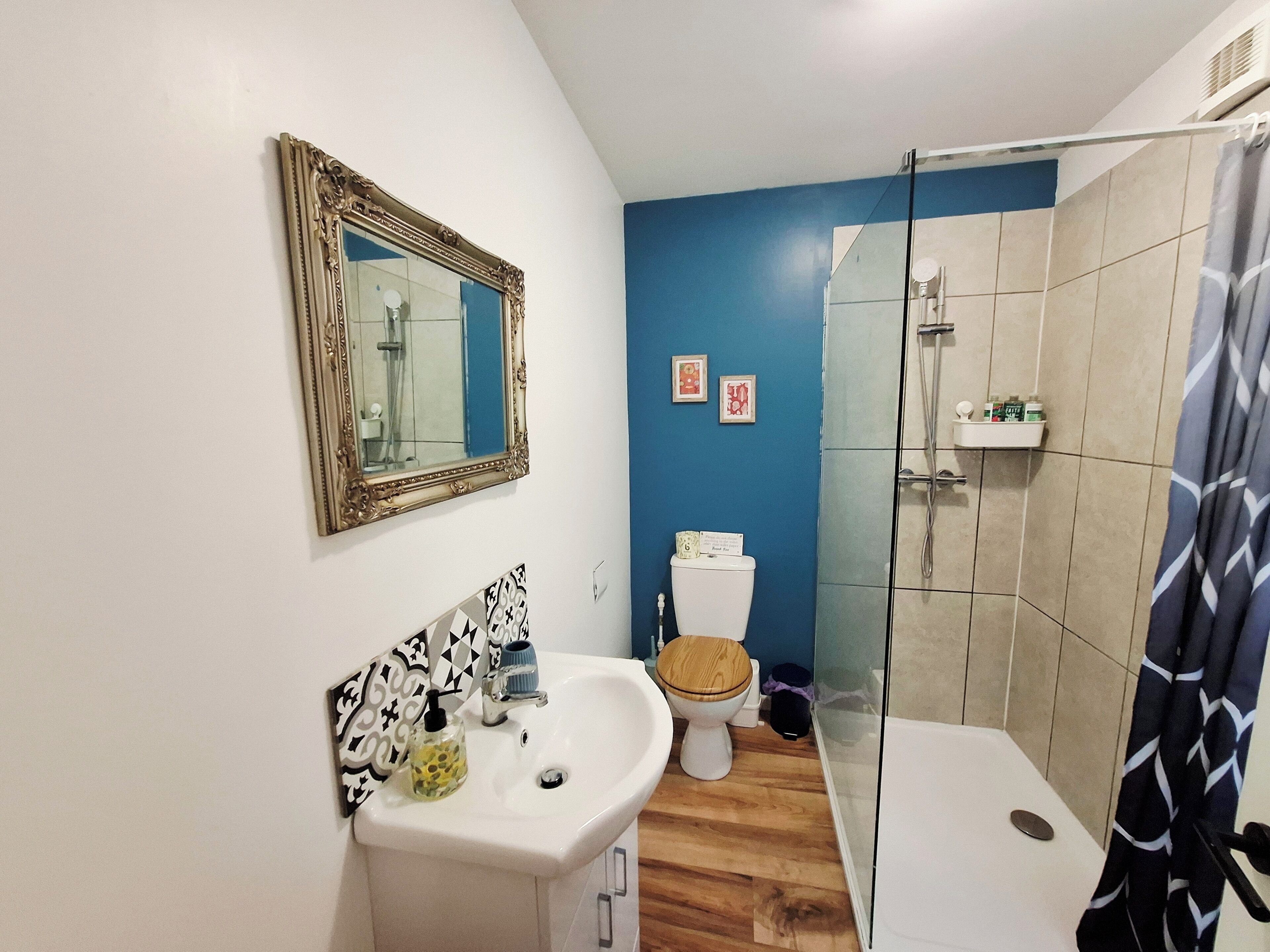 Une salle de bain avec un lavabo blanc, des toilettes avec un abattant en bois, une douche avec une porte en verre et un mur bleu.