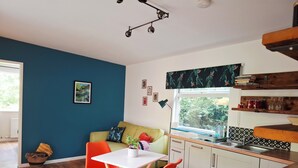 A modern kitchen with a dining area, featuring a white table, orange chairs, and a blue wall. - The Hide - A Secret Cabin in the Trees: Peace Nature & Privacy! (Tiverton)