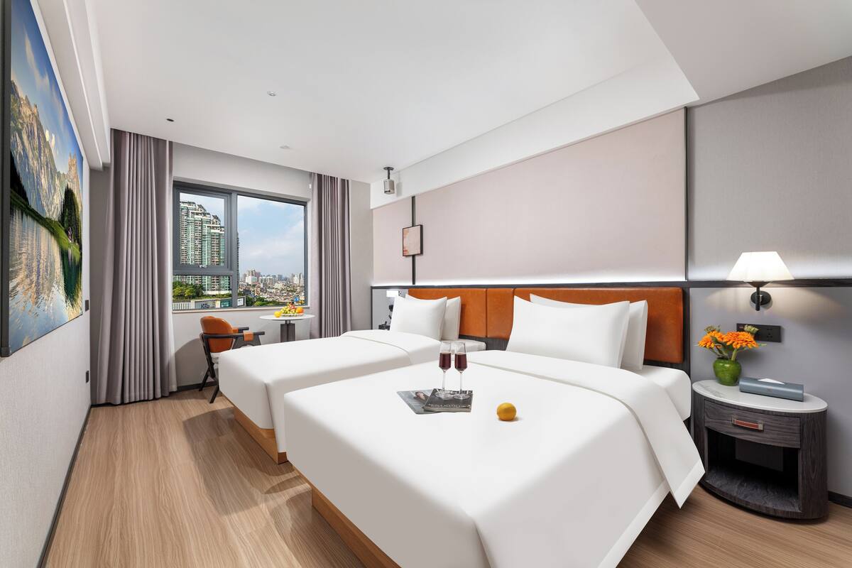 A hotel room with two beds, a city view, a small table with a lamp, and a painting on the wall.