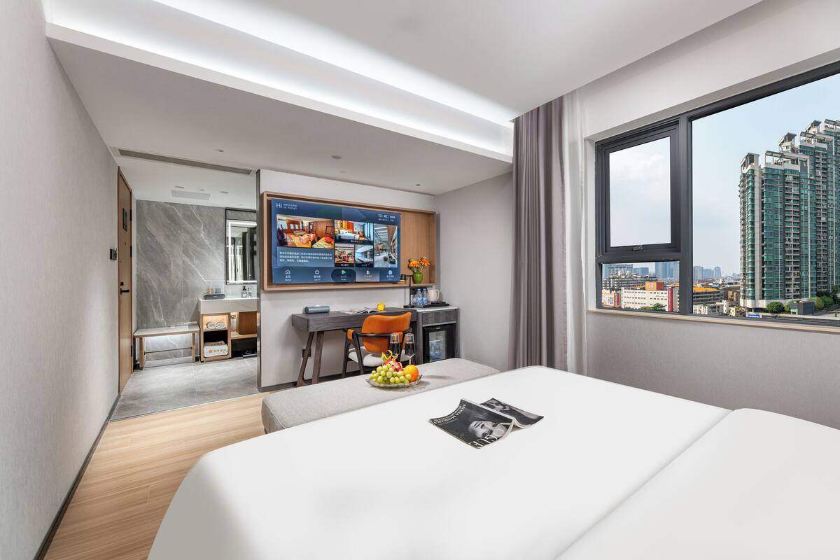 A modern hotel room with a bed, desk, and chair, a large window with a city view, and a digital screen displaying room amenities.
