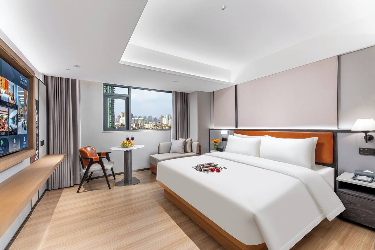 A modern hotel room with a large bed, a desk with a chair, a small table with a fruit bowl, and a city view through the window.
