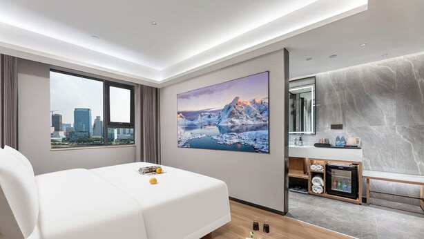 Habitación de hotel moderna con una cama grande, vistas a la ciudad, un cuadro de un paisaje montañoso, un minibar pequeño y un banco de madera.