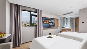 Habitación de hotel con una cama grande, un escritorio con fruta, un televisor mostrando los servicios de la habitación y vistas a la ciudad.