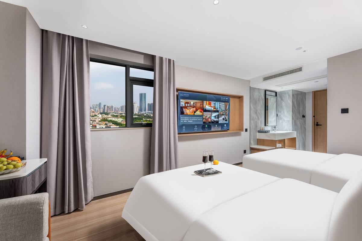 A hotel room with a large bed, a desk with fruit, a TV displaying room amenities, and a city view.