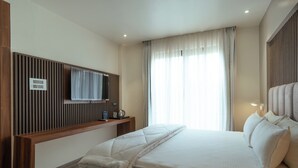 Une chambre d’hôtel moderne dotée d’un grand lit, d’un téléviseur à écran plat fixé sur un panneau en bois, d’une table de chevet avec une lampe et d’une fenêtre ornée de rideaux légers.