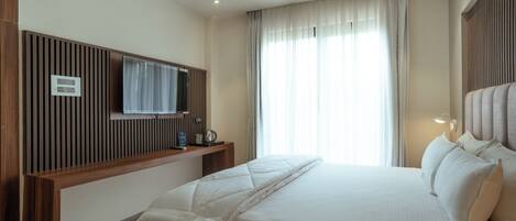 A modern hotel room with a large bed, a flat-screen TV mounted on a wooden panel, a bedside table with a lamp, and a window with sheer curtains.
