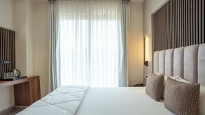 Une chambre d’hôtel avec un grand lit, une console en bois, une lampe de chevet et une vue sur l’extérieur à travers des rideaux transparents.