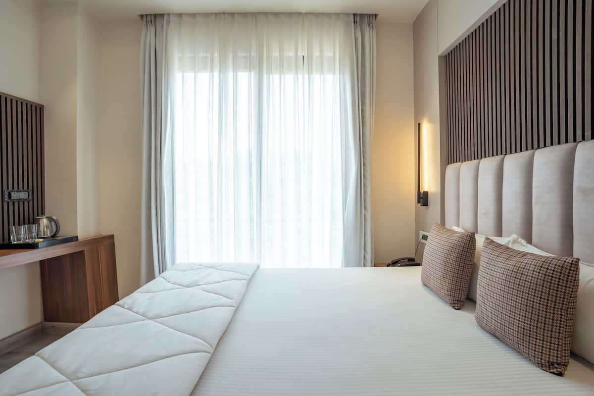 Une chambre d’hôtel avec un grand lit, une console en bois, une lampe de chevet et une vue sur l’extérieur à travers des rideaux transparents.