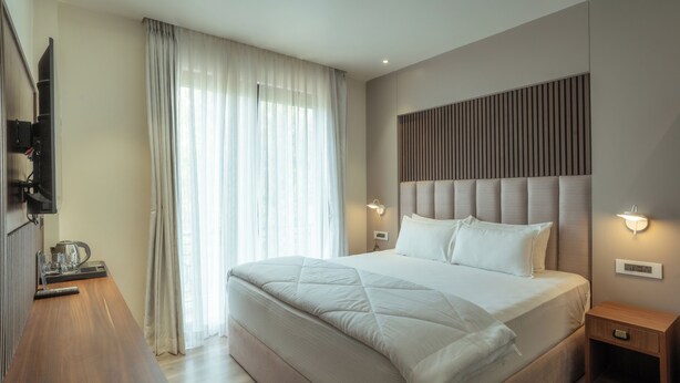 Une chambre moderne avec un grand lit, une table de chevet en bois, un téléviseur à écran plat fixé au mur et une fenêtre ornée de rideaux légers.
