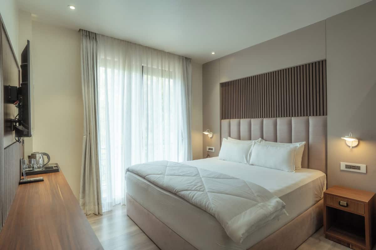 Une chambre moderne avec un grand lit, une table de chevet en bois, un téléviseur à écran plat fixé au mur et une fenêtre ornée de rideaux légers.