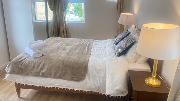 Een net opgemaakt bed met een witte deken, een grijze plaid en twee blauwe kussens. Er staat een nachtkastje met een lamp en een klein tafellampje.
