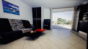 Ein Wohnzimmer mit einer schwarzen Couch, einem roten Couchtisch, einem Flachbildfernseher und einem großen Fenster mit Blick ins Grüne.