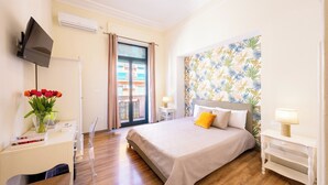 Un dormitorio con cama, escritorio con televisor, ventana con cortinas y pared con papel tapiz tropical.