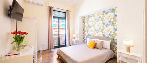 Un dormitorio con cama, escritorio con televisor, ventana con cortinas y pared con papel tapiz tropical.
