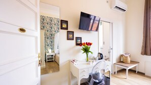 Habitación de hotel con escritorio blanco, una silla, televisor montado en la pared y una mesita con una cesta.