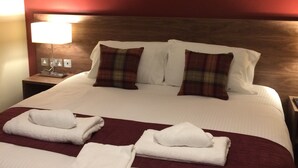 A neatly made bed with white linens, two plaid pillows, and two folded towels on a red bedspread. There are two framed pictures on the wall above the bed.