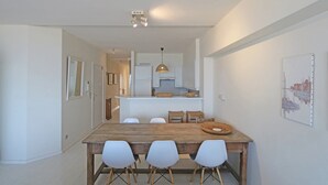 Ein Essbereich mit Holztisch, vier weißen Stühlen, einer Küche im Hintergrund und einer Pendelleuchte über dem Tisch.