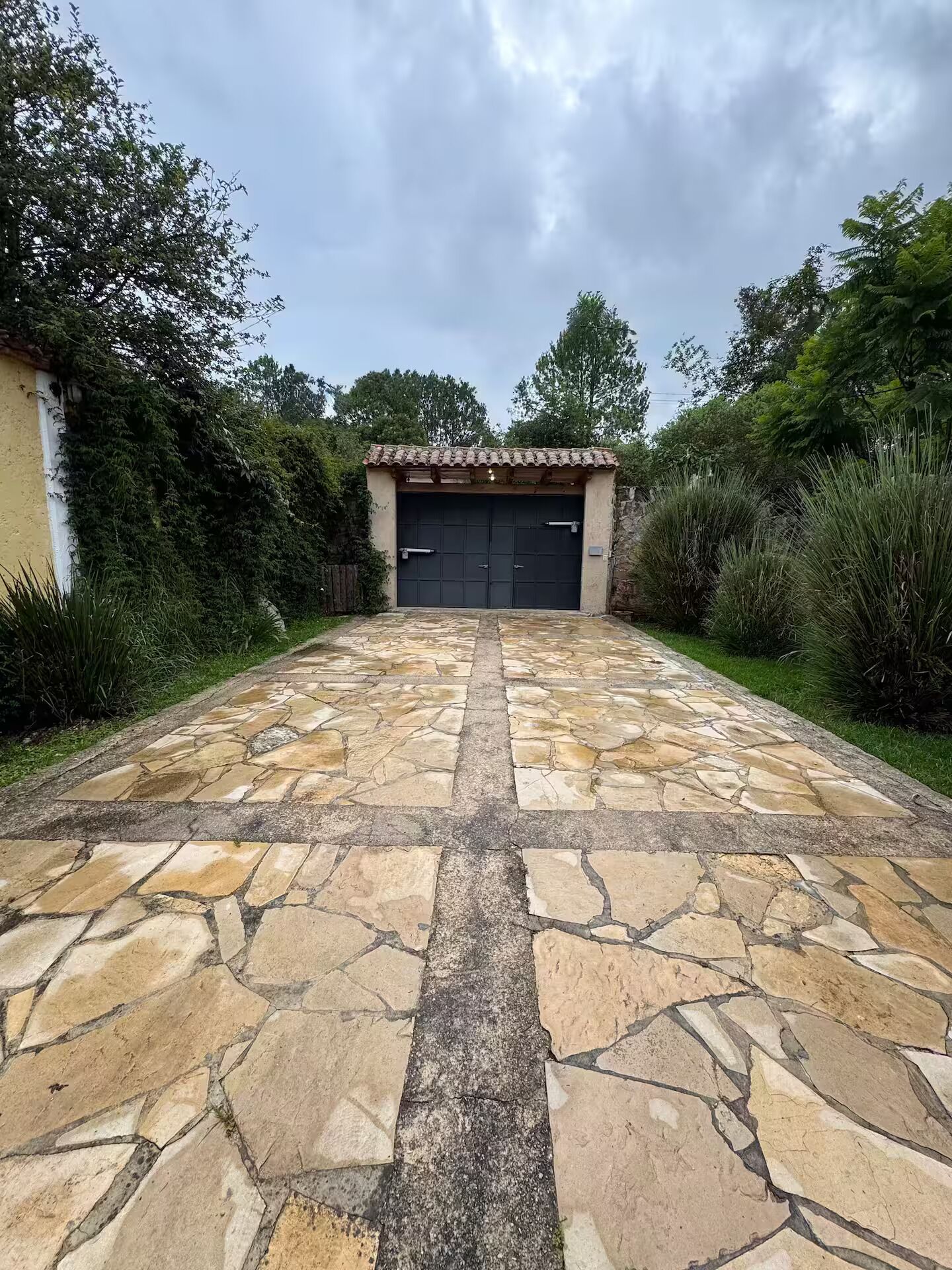Une allée dallée de pierres menant à un garage surmonté d’un toit en tuiles, le tout entouré de verdure.
