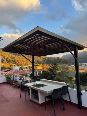 A covered outdoor seating area with a table and chairs, overlooking a scenic landscape with trees and buildings.