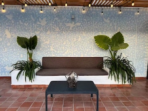 Une pièce avec un mur orné de motifs, un canapé marron, deux grandes plantes en pot, une petite table sur laquelle est posé un objet décoratif, et un sol carrelé.