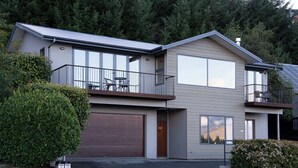 Une maison moderne à deux étages, dotée d’un garage, d’un balcon avec mobilier d’extérieur et offrant une vue sur la colline boisée.