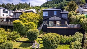 A modern house with a large deck, surrounded by well-maintained gardens and a forested hillside.