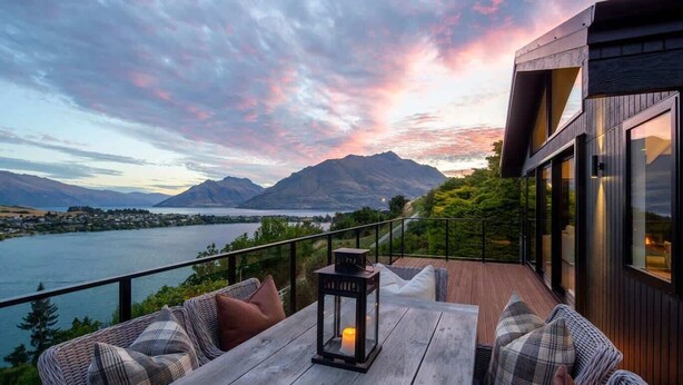 A wooden deck with a table and chairs, overlooking a lake and mountains.