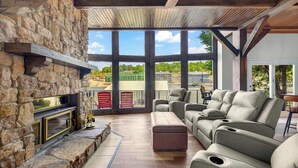 A stone fireplace, a large window, and a seating area with a view of buildings and trees. - Cozy in a Cove – 14 ppl Stay (Camdenton)