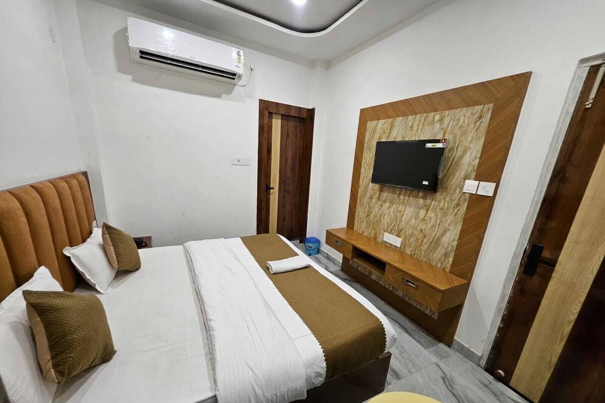 A hotel room with a bed, a TV mounted on a wooden wall, a small table, and an air conditioning unit.